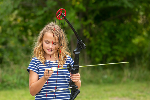 Front view of a young girl practicing archery at an outdoor shooting range on an early autumn day in Minnesota, USA. In this image, the girl is loading the arrow onto her bow to get ready to shoot at a target.