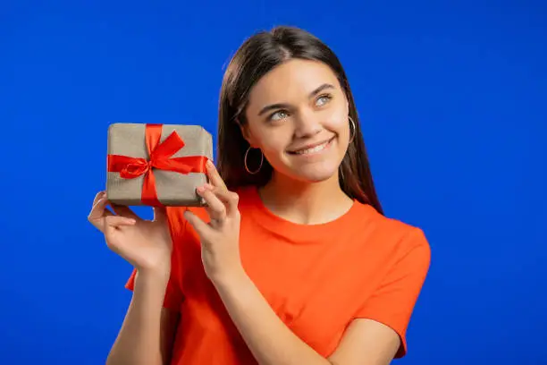 Excited woman received gift box with bow. She is happy and flattered by attention. Girl smiling with present on blue background. Studio portrait.