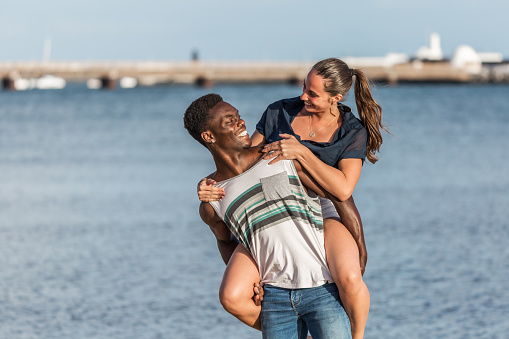 African American male smiling and looking at happy female while carrying girlfriend on back near sea on Lanzarote island, Spain