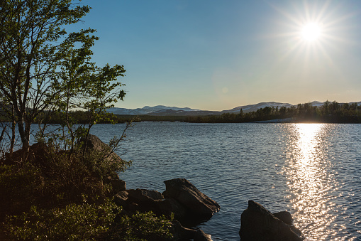 Summer view across a high lake in the northern Swedish mountains. Snow covered mountain tops in the distance and a bright sun with sun rays in the clear blue sky