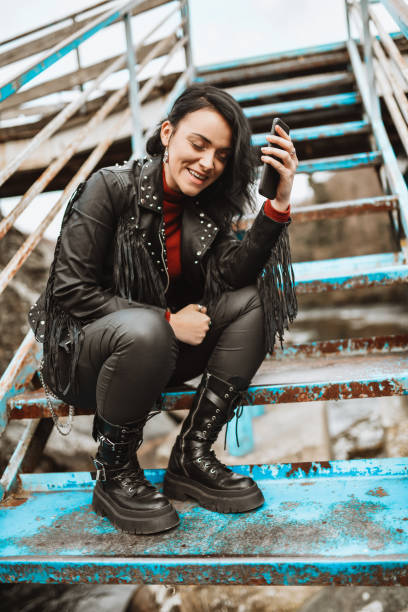 donna punk sorridente che usa lo smartphone mentre è seduta su una scala di metallo all'esterno - rock staircases foto e immagini stock