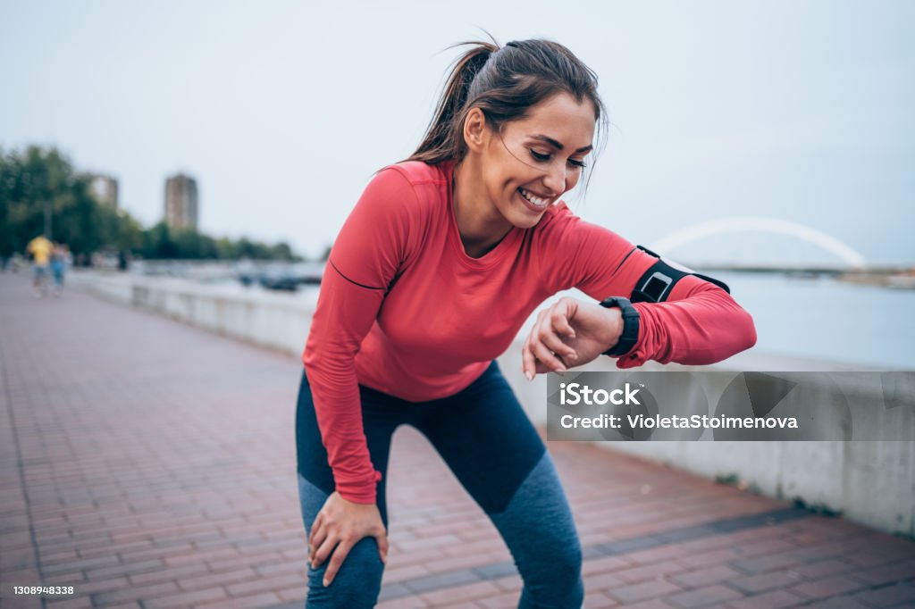 Sporty young woman checking the time after jogging. Shot of a beautiful smiling young sportswoman resting after workout outdoors. Young woman checking pulse on her smart watch. Smart Watch Stock Photo