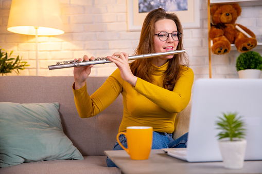 Young woman in yellow pullover showing how to play flute via white laptop. Side hustle job as a music teacher online, working on weekends. Online music tuition.