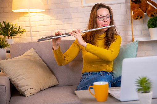 Young woman in yellow pullover playing flute via white laptop. Practicing music instrument online. Side hustle job as a music teacher online, working on weekends. Online music tuition.
