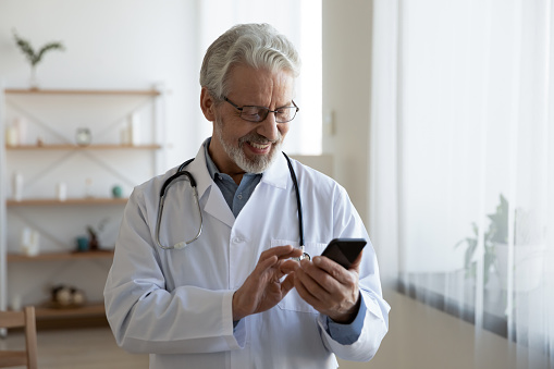 Smiling mature Caucasian male doctor or GP use cellphone browse internet in modern hospital. Happy senior man therapist look at smartphone screen consult clinic patient online on device.