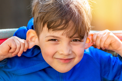 Caucasian blond Smiling boy pulling stretching his ears looking at the camera sitting at his home backyard, is a backlit headshot taking outdoors