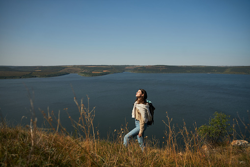 Attractive young lady in casual wear hiking with backpack among beautiful nature near Dniester river. Scenic landscape on background.