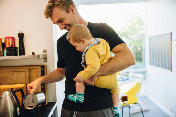 Man on paternity leave Father carrying his baby and preparing coffee in kitchen. Man on paternity leave with his toddler standing in kitchen and making coffee. paternity stock pictures, royalty-free photos & images