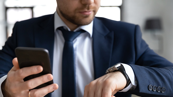 Close up confident businessman wearing suit holding smartphone, checking time, looking at wrist watch on hand, planning workday, waiting for meeting, appointment, task management concept