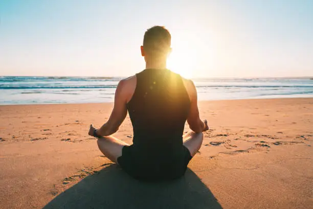 Photo of Healthy man practicing yoga and meditates on the beach with ocean view at sunset or sunrise. Sitting in easy pose or sukhasana with mudra on the sand. Relaxation, harmony, freedom. Healthy lifestyle