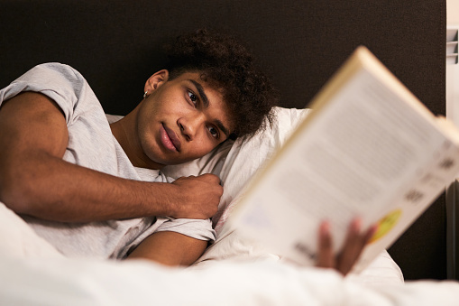 Avoid gadgets before bed. Portrait of calm brunette guy lying in the bed at home, resting and reading interesting book before going to sleep. Leisure, relaxation concept