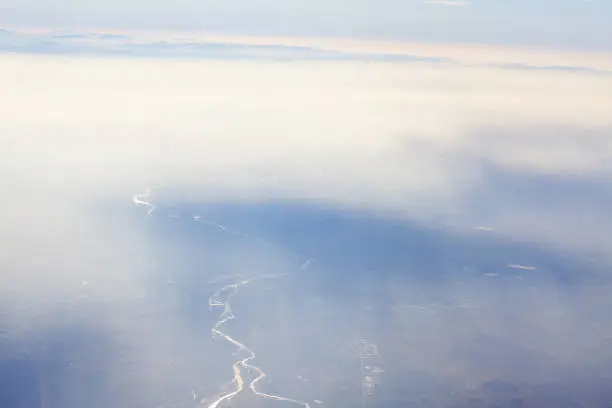 The earth is seen through transparent clouds . Aerial view of river meanders and haze . Fog with sun rays over the earth