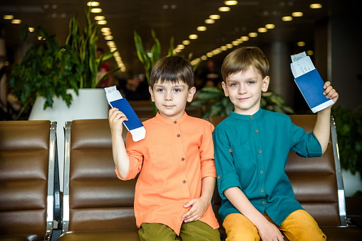 Two young kids sitting on brown chairs, holding passports and tickets in waiting hall in airport. Travel and holidays with children concept.