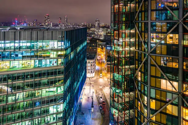 High angle view of highly detailed and illuminated modern office building during night hours in London City, UK.  Downtown street with unrecognisable people and traffic with Canary Wharf in the far background. Photographed on Canon EOS R full frame system.