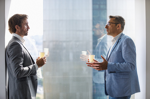 Man standing by the floor to ceiling window on coffee break and seriously discussing business with colleague