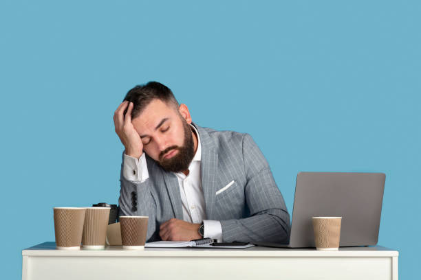istock Exhausted businessman surrounded by empty coffee cups sleeping at workplace, blue studio background 1309037877