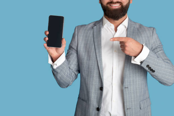 istock Unrecognizable young businessman pointing at smartphone with empty screen on blue background, mockup for design 1309038016