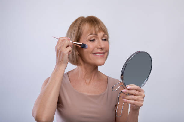 istock Lovely senior lady using cosmetic brush to apply decorative makeup on her face, looking in mirror on light background 1309039356