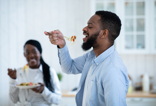 Cheerful Black Man Tasting Spaghetti While Having Lunch With Wife In Kitchen, Romantic African American Couple Enjoying Cooking And Eating Tasty Food At Home, Selective Focus On Guy
