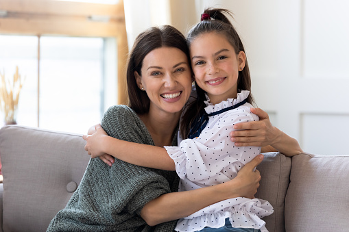 Portrait of happy young Caucasian single mom hug cuddle small schoolgirl daughter look at camera posing smiling. Overjoyed mother and little girl child embrace relax together show love and care.