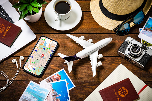 Top view of an tiny airplane surrounded by various traveling stuff such as a coffee cup, a camera with some snapshots, a smartphone with a map on the screen, an opened notebook, two passports with boarding passes and some summer accessories like a hat and sunglasses. All the objects are on a rustic wooden desk. Studio shot taken with Canon EOS 6D Mark II and Canon EF 24-105 mm f/4L