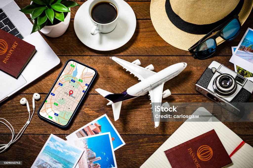 Travel planning background Top view of an tiny airplane surrounded by various traveling stuff such as a coffee cup, a camera with some snapshots, a smartphone with a map on the screen, an opened notebook, two passports with boarding passes and some summer accessories like a hat and sunglasses. All the objects are on a rustic wooden desk. Studio shot taken with Canon EOS 6D Mark II and Canon EF 24-105 mm f/4L Travel Stock Photo