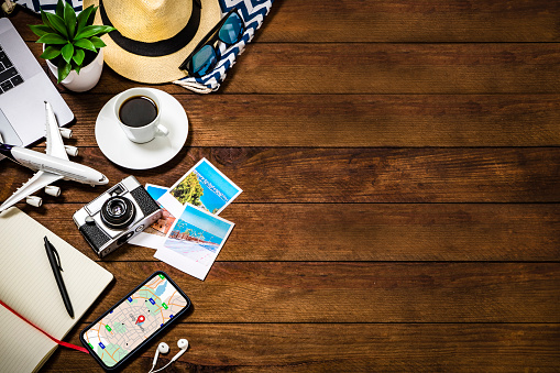 Top view of various traveling stuff such as an opened notebook, a coffee cup, a camera with some snapshots, a smartphone with a map on the screen, a tiny airplane, and some summer accessories like a hat and sunglasses. All the objects are at the left side of the image leaving a useful copy space at the right side on a rustic wooden desk.  Studio shot taken with Canon EOS 6D Mark II and Canon EF 24-105 mm f/4L