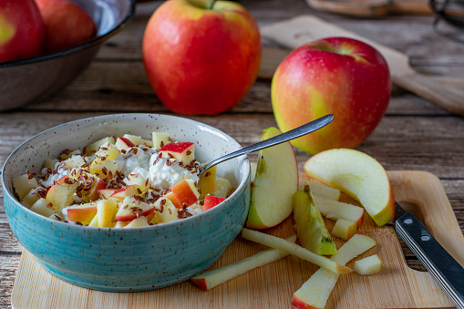 Still life of a healthy breakfast bowl with cottage cheese, apples and linseeds served on a wooden cutting board on kitchen table from above.