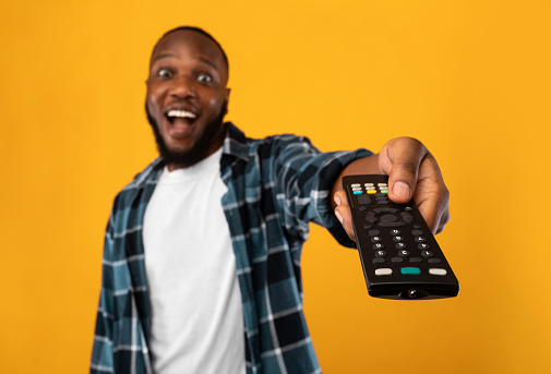 Excited African American Man Pointing Television Controller To Camera Switching Channels Watching TV Standing Over Yellow Studio Background. Television Programming Advertisement. Selective Focus