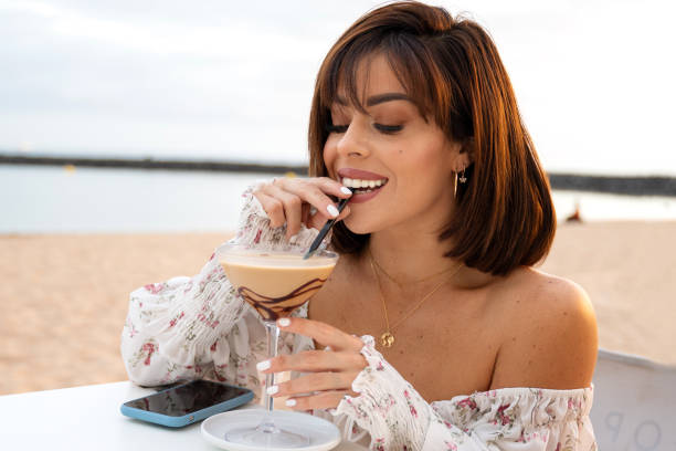 alegre chica morena de moda bebiendo café helado en el bar de la playa, relajándose, usando el teléfono móvil y sonriendo. - bangs fotografías e imágenes de stock