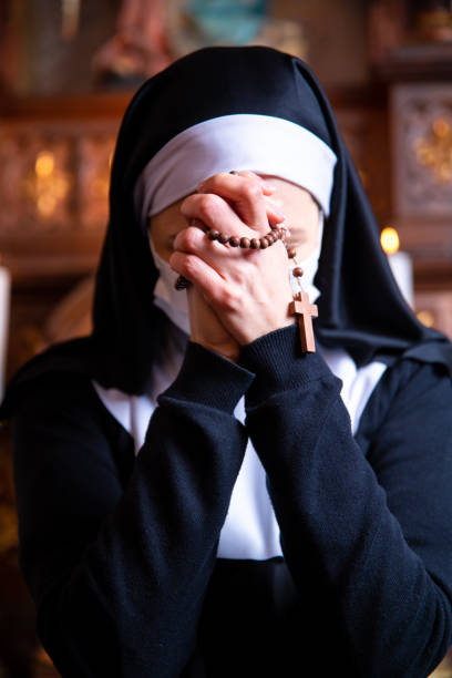 Nun praying in church, wearing protective face mask. Close up on hands holding rosary beads. Nun holding hands together, praying to God, unrecognizable face. She is wearing a white surgical face mask. A nun bowing her head in prayer. nun praying cross human hand stock pictures, royalty-free photos & images