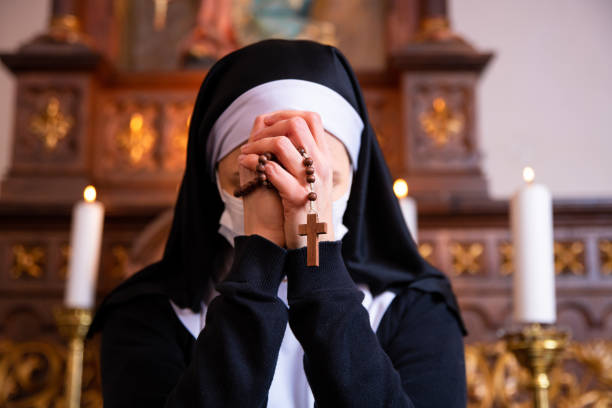 A nun bowing her head in prayer. Close up on hands holding rosary beads. Nun holding hands together, praying to God, unrecognizable face. She is wearing a white surgical face mask. nun praying cross human hand stock pictures, royalty-free photos & images