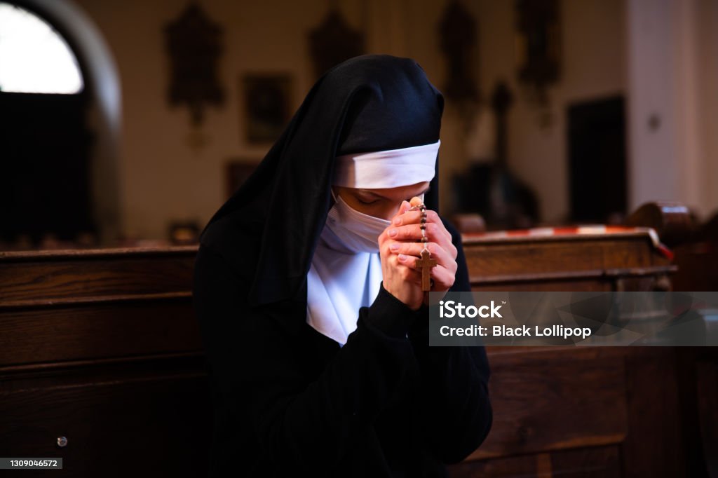 A nun bowing her head in prayer sitting in a church wearing protective face mask. Nun holding hands together, praying to God, unrecognizable face. She is wearing a white surgical face mask. Nun Stock Photo