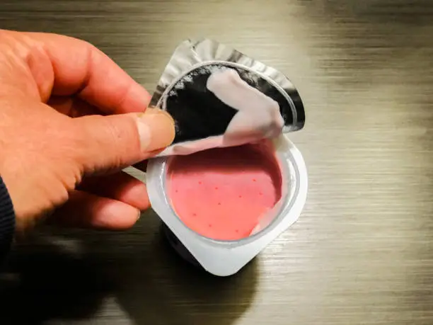 A BPA-free plastic container with strawberry yogurt. A hand is opening the foil wrap lid of the container.  There is copy space to the right of the image.