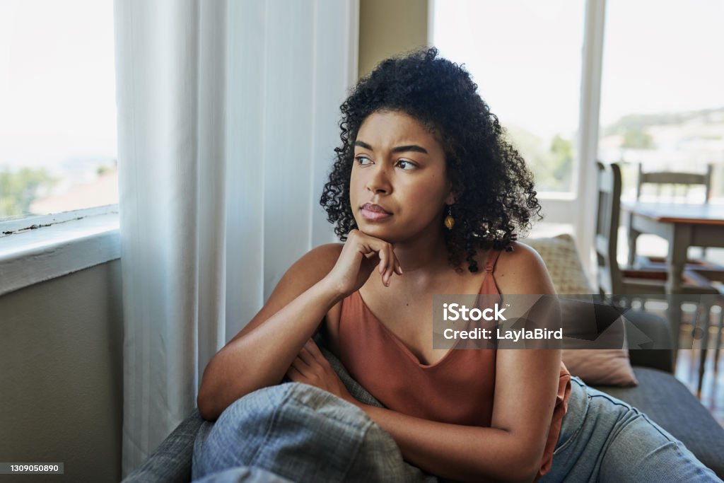 Sitting with the worries of an uncertain future Shot of a young woman looking pensively out a window at home Women Stock Photo
