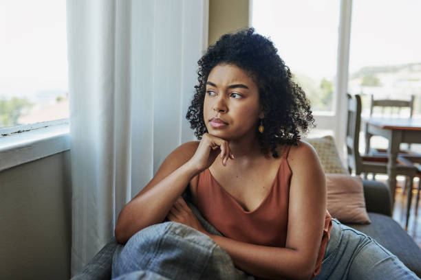Sitting with the worries of an uncertain future Shot of a young woman looking pensively out a window at home sadness stock pictures, royalty-free photos & images