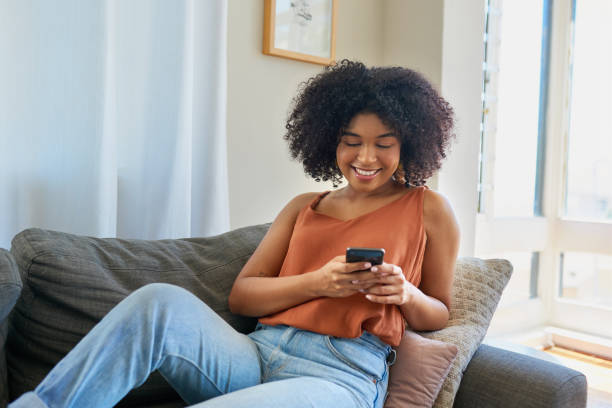 Clocking high scores on her favourite mobile games Shot of a young woman using a cellphone while relaxing on a sofa at home scrolling photos stock pictures, royalty-free photos & images
