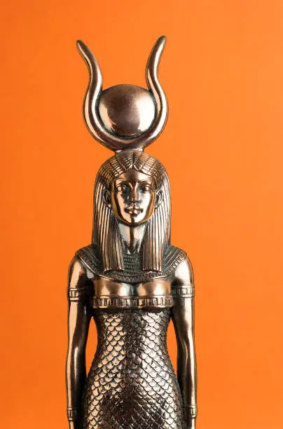 The figure of the Egyptian goddess Isis on an orange background. Bronze statuette of the mother of fertility.
