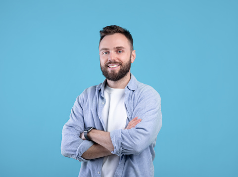 Portrait of positive young guy posing with crossed arms and smiling at camera on blue studio background. Masculine bearded millennial man in casual clothes having happy face expression