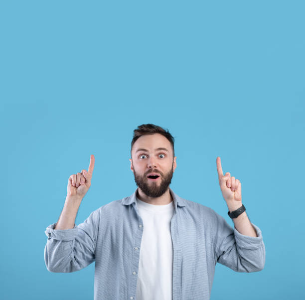 istock Handsome bearded guy having shocked face expression, pointing at blank space over his head on blue studio background 1309055097