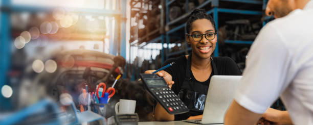 sell customer service and cheerful seller, African woman store show prize on calculator, explain quality and product insurance to client, service mind and happy cashier smiled in auto parts for sale stock photo