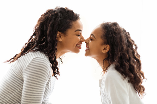 African american mom and daughter touching each other with noses over white background, side view. Happy black young woman and school girl mother and child bonding over white