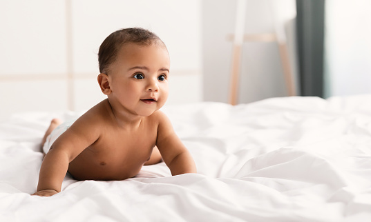 Portrait of a cute little African American baby lying on the white blanket at home. Black naked infant child crawling on the bed in the bedroom. Free copy space, selective focus, blurred background