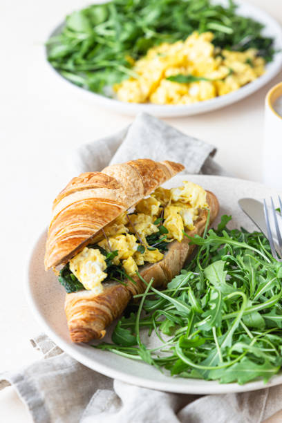 istock Delicious croissant sandwich with scrambled spinach eggs served with arugula on plate and coffee cup, light concrete background. 1309057173