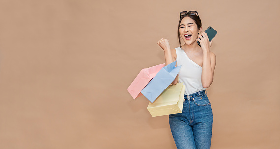 Portrait of excited beautiful asian girl with fashion sunglasses holding shopping bags screaming isolated on orange background. Happy beautiful asian shopaholic woman carrying shopping bags with copy space