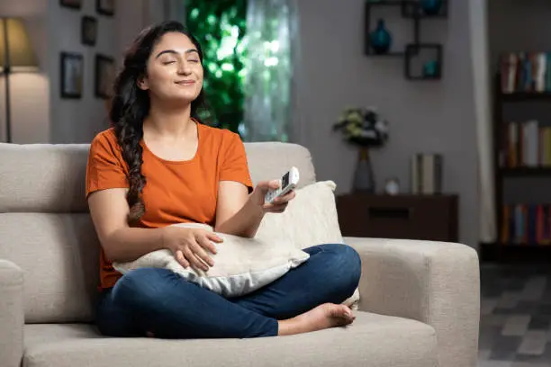 Photo of shot of a young women Turning on the air conditioner sitting on sofa at home:- stock photo
