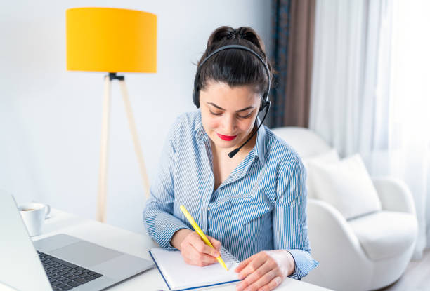 istock Smiling young woman with headset doing video call at home office. Study using computer and write in notebook. Remote work from home 1309071592