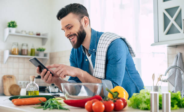 istock Happy handsome bearded man is using his smart phone while he preparing vegan healthy breakfast for a lovely family 1309072978