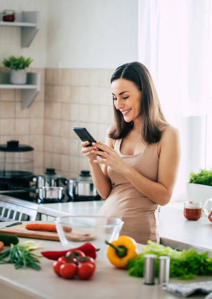 nowoczesna szczęśliwa młoda urocza kobieta relaksująca się podczas korzystania z inteligentnego telefonu w kuchni w domu - vegan food cheerful vertical indoors zdjęcia i obrazy z banku zdjęć