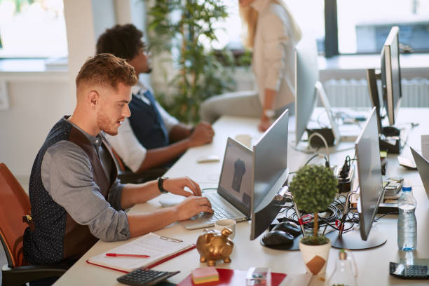 young designer typing on computer keyboard at workplace in the open space office with colleagues in the background. casual, business agency